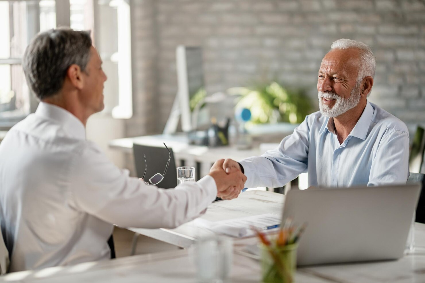 Two smiling men shake hands while seated in a business meeting.